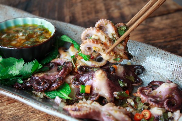 Grilled baby octopus on a long gray serving platter with a small bowl of dipping sauce on the side and a pair of chopsticks lifting up a baby octopus.