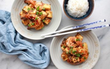 Two white bowls of General Tso's Shrimp and Tofu with chopsticks and a bowls of white rice on the side along with a blue napkin.
