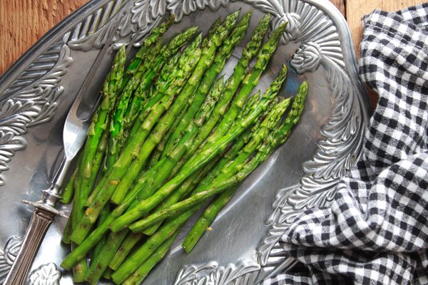 Vibrant grilled asparagus spears on a silver serving platter with a silver asparagus fork along side with a black and white checkered napkin on the side.