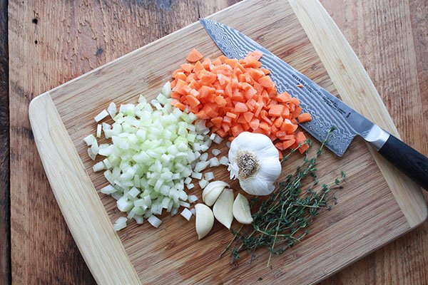 A mirepoix of chopped vegetables on a wooden cutting board with a chef's knife on the side.