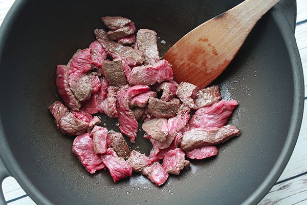 Sliced sirloin beef being stir-fried in a large wok with a wooden spoon inserted.