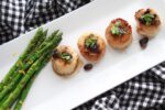 Beautifully plated seared sea scallops drizzled with a black bean sauce with grilled asparagus on the side on top of a long white plate beneath a black and white checkered napkin.