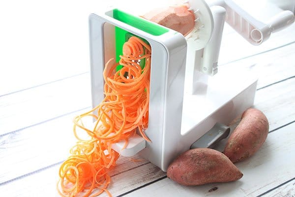 A large white spiralizer spiralizing a sweet potato with two whole potatoes on the side on top of a white plank board.