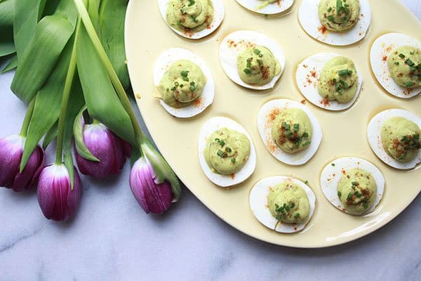 A yellow egg platter filled with avocado deviled eggs, topped with chili powder and chives and a bunch of purple tulips on the side.