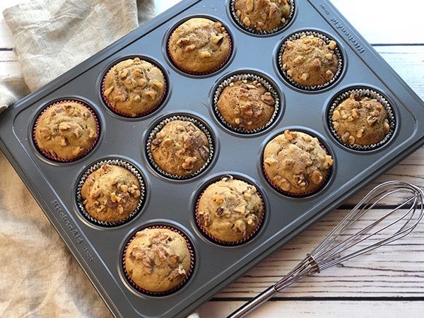 Banana nut muffins baked in a muffin tin right out of the oven on top of a white wooden board with a whisk on the side.