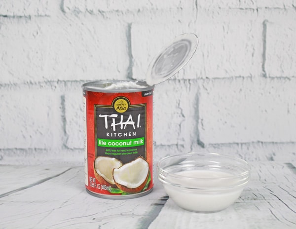A can of coconut milk and a small glass bowl filled with coconut milk placed on top of a white plank board with white brick in the background.