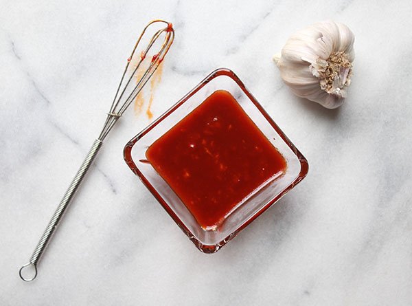 A small square glass bowl filled Gochujang sauce and a small whisk and garlic bulb on the side on top of a marble surface.
