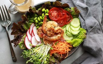 Grilled miso shrimp on top of greens with cucumbers, tomatoes, edamame pods, radishes and carrots with a grey napkin and silver fork on the side and miso dressing in a clear glass in the background.
