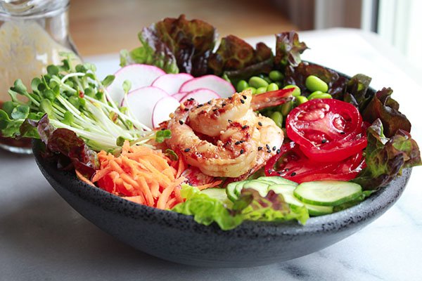 Grilled miso shrimp on top of greens with cucumbers, tomatoes, edamame pods, radishes and carrots on top of a marble surface and miso dressing in a clear glass in the background.