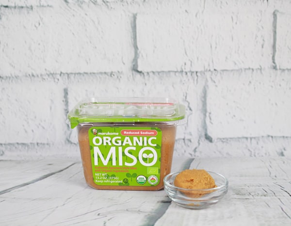 A plastic container of organic miso paste and a small glass bowl filled with miso paste placed on top of a white plank board with white brick in the background.