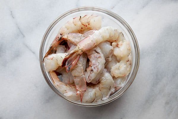 A clear glass bowl filled with pieces of peeled pink shrimp, tail on, on top of a marble board.