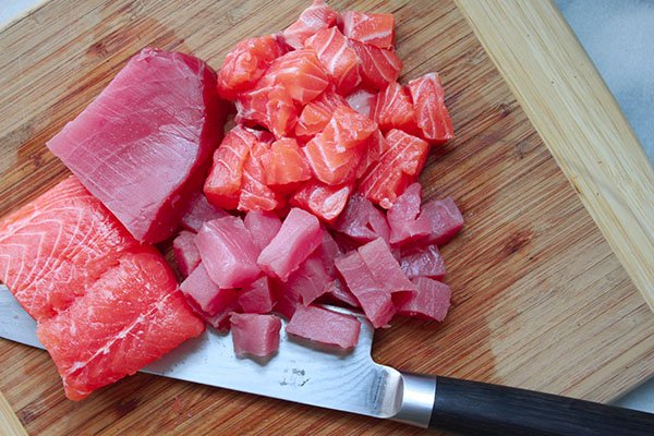 Cubes of sushi-grade salmon and tuna on top of a wooden cutting board with a chef's knife on the sidel