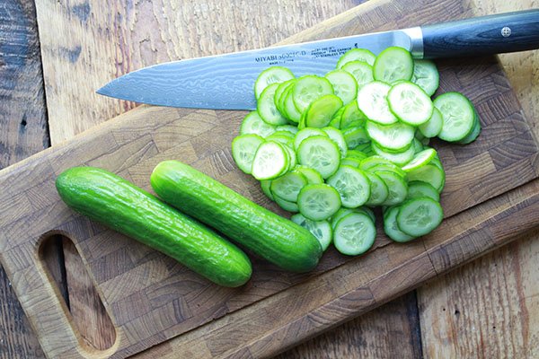 Thinly sliced Persian cucumbers in a pile on top of a wooden cutting board with two uncut cucumbers and a chef's knife on the side.