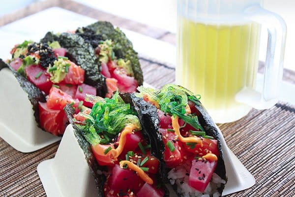 Beautiful sushi-grade tuna and salmon cubes nestled inside a nori shell on top of white rice and topped with black caviar and wakame seaweed salad placed in taco holders on top of a bamboo placemat with a frosty mug of beer in the background.