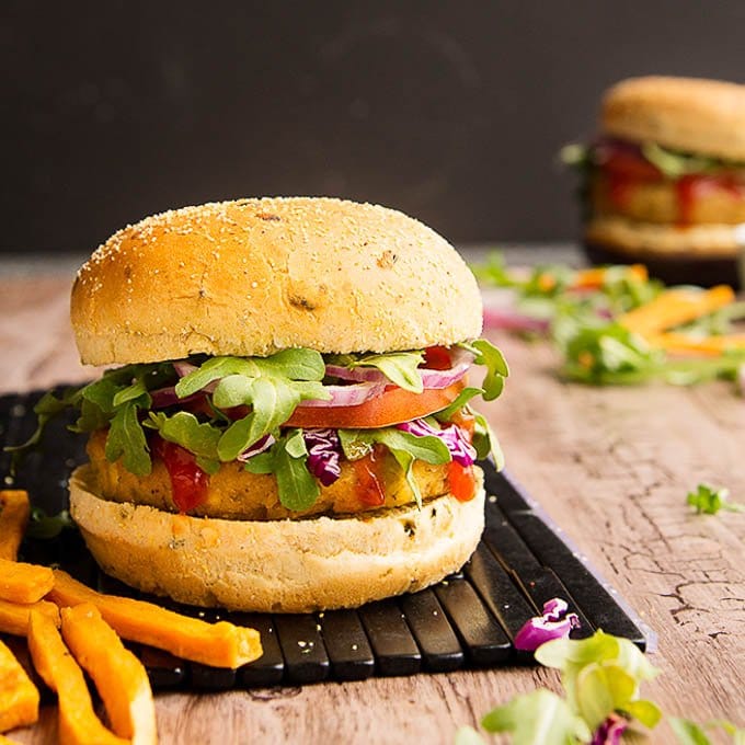 A thick vegetarian tofu burger sandwiched in between a burger bun and topped with greens, tomatoes, and red onions on top of a black plank board with fries on the side.