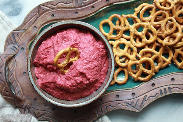 Vibrant red beet hummus in a gray bowl with a side of pretzels for dipping on a green serving bowl.