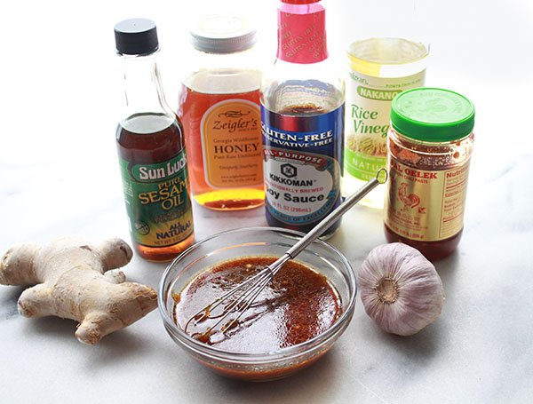 Asian sauce ingredients, gluten-free soy sauce, sesame oil, sambal oelek, rice vinegar, honey, and fresh ginger and garlic on a marble surface.
