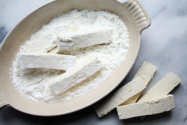tofu strips being dredged in cornstarch inside an oval dish placed on top of a marble surface