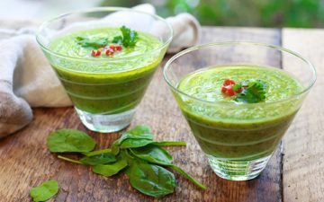 green gazpacho soup in two clear class bowls with fresh spinach on this side on top of a wooden board