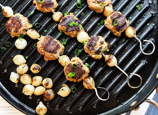 Metal skewers of grilled meatballs with pearl onions in between on top of a grill pan and grilled pearl onions on the side.