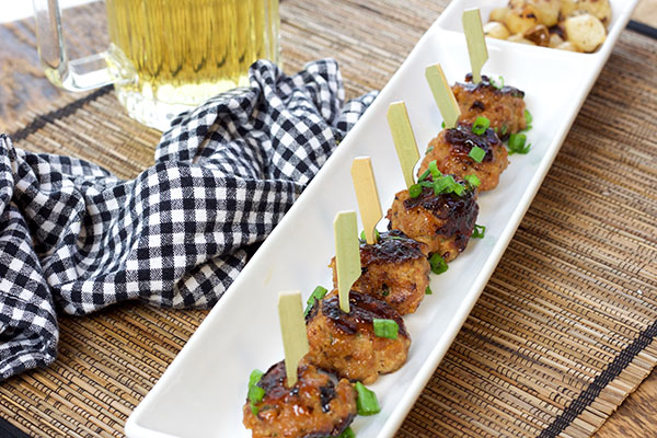 A row of skewered sweet and spicy BBQ meatballs on top of a thin white rectangular serving platter with a black and white checkered napkin and mug of beer on the side.