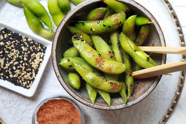 A bowl of spicy edamame pods topped with sesame seeds and sriracha seasoning on the side.