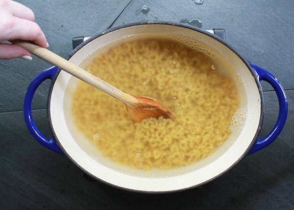 A woman's hand stirring elbow noodles in boiling water with a wooden spoon inside a large blue Dutch oven on top of a black surface.