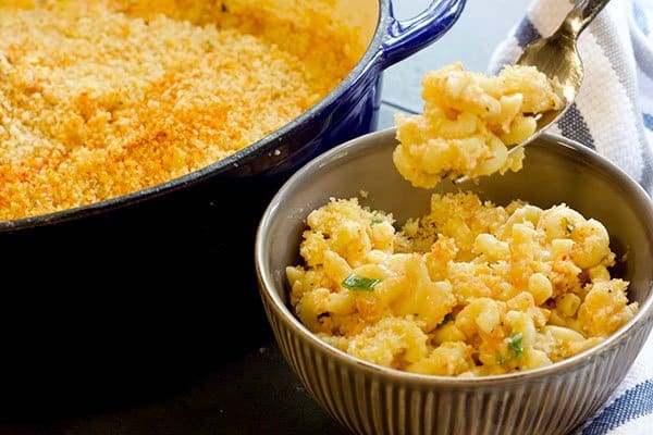 A fork holding a bite-sized portion of Kimchi mac and cheese inside a gray bowl with a large blue Dutch oven filled with baked mac and cheese.