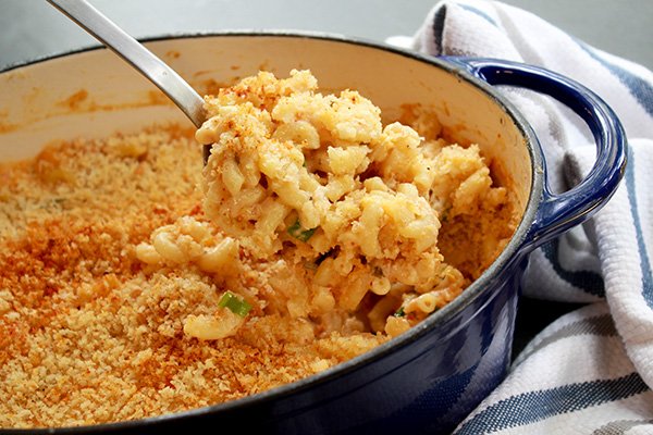 A large blue Dutch oven filled with creamy kimchi mac and cheese with a serving spoon scooping some out and a striped kitchen towel on the side.