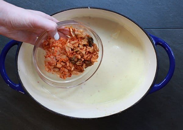 A woman pouring kimchi from a glass bowl into a blue Dutch oven filled with melted creamy cheese on top of a black surface.