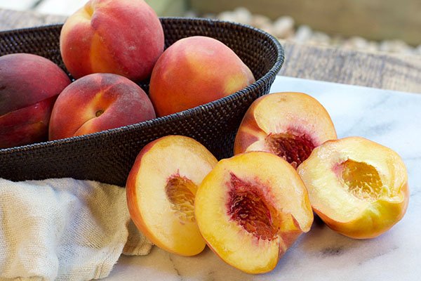 fresh peaches halved and whole peaches in a wicker bowl on top of a marble surface