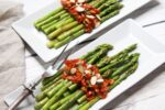 Beautiful grilled asparagus spears with an Asian sweet and spicy pepper sauce placed on top of two narrow white plates with an asparagus spear on top.