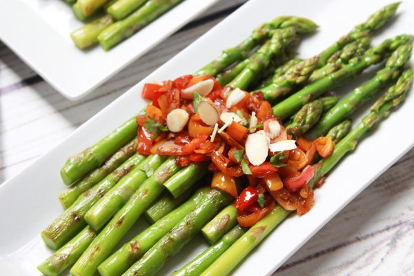 Vibrant grilled asparagus spears on a narrow white plate topped with a roasted pepper and almond sauce.