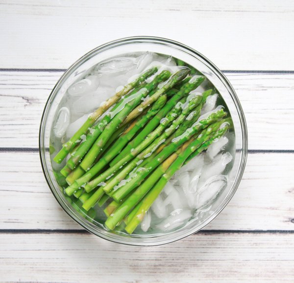 Blanched asparagus spears bathing in an ice bath inside a glass bowl on top of a white plank board.