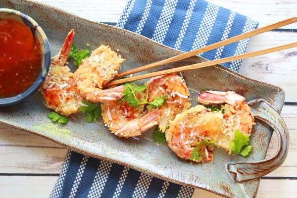 Baked coconut shrimp on a gray serving platter with wooden chopsticks on top and a small bowl of chili sauce on the side.