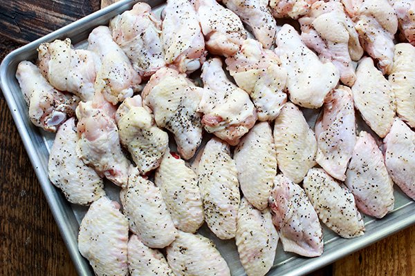 Three rows of raw chicken wings spiced with black pepper on a stainless baking sheet.