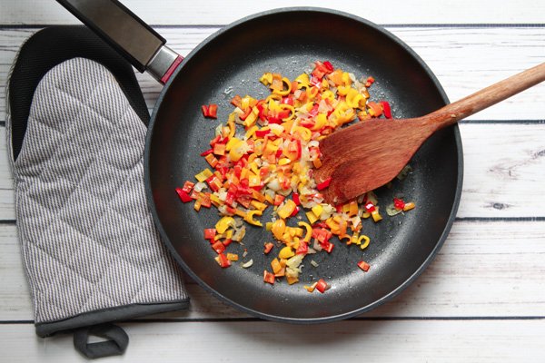Multi-colored bell peppers being sauteed in a frying pan with a wooden spatula and an oven mitt on the side on top of a white plank wooden board.