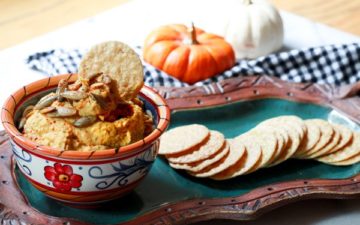A small dipping bowl filled with pumpkin hummus topped with pumpkin seeds served on top of a green platter with rice crackers on the side and a black and white checkered napkin on the side.