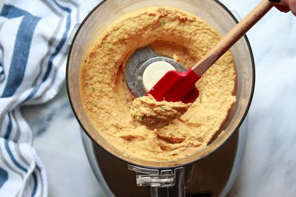 A food processor with pureed pumpkin hummus inside with a red rubber spatula inserted place on top of a marble surface and a kitchen towel on the side.