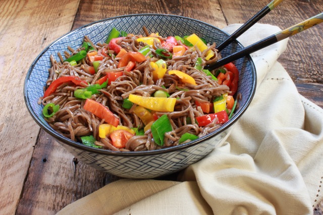 Colorful Japanese soba noodle salad in a large blue bowl with chopsticks on top of a wooden board and a napkin on the side