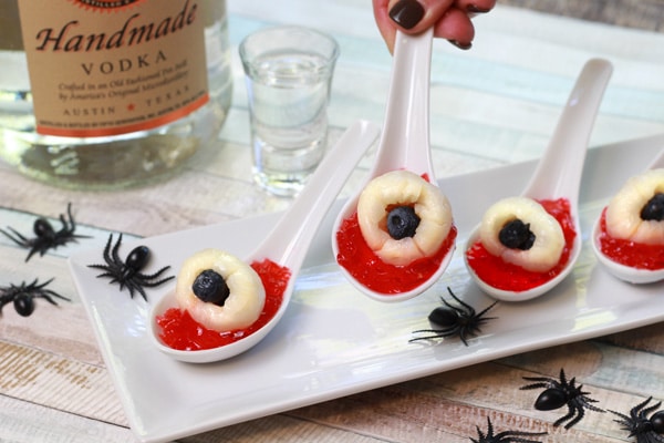 eyeball jello shots on small white spoons on top of a white platter with black spiders sprinkled around and a bottle of Vodka in the background