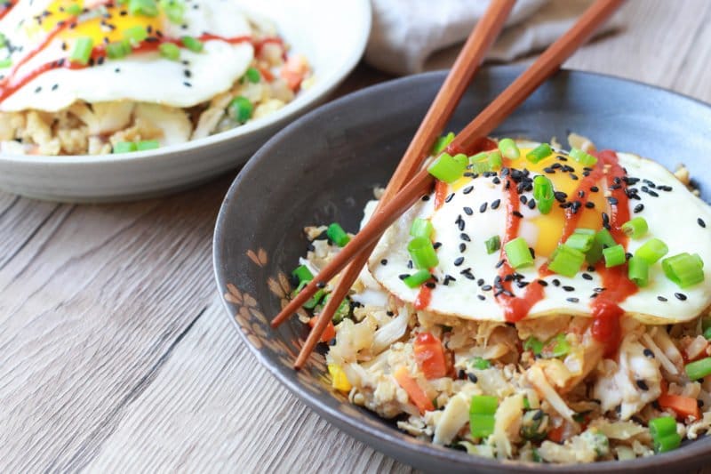 A gray bowl filled with cauliflower fried rice with crab topped with a fried egg and a pair of chopsticks on top of a wooden board with another bowl of fried rice in the background.