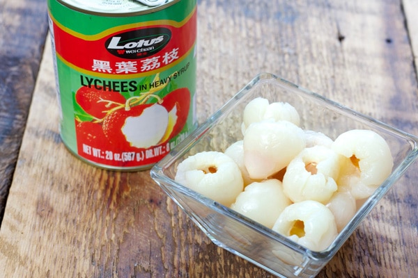a clear bowl of lychees with a can of lychees along side on a wooden board