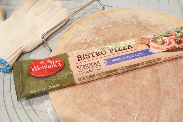 A package of Bistro Pizza thin crust pizza dough sitting on top of a pizza stone with a white oven mitt on the side.
