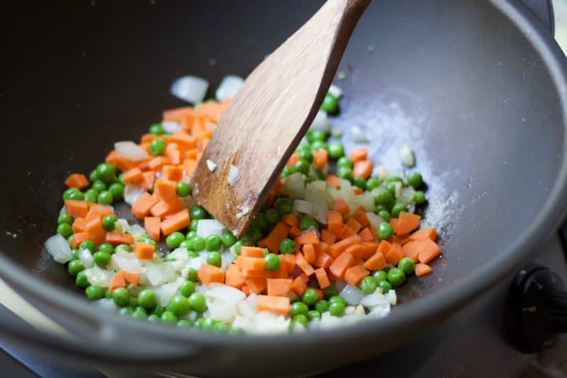 Chopped carrots, onions, and peas being stir-fried in a large wok with a wooden spatula.