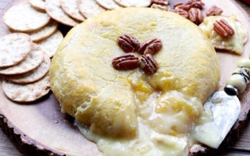 A golden-brown baked brie oozing with cheese and topped with pecans right out of the oven placed on top of a wooden board with crackers and pecans placed behind.