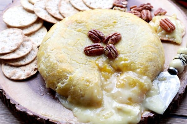 a golden-brown round baked brie in puff pastry with cheese oozing out on top of a wooden serving plate with walnuts on top and crackers on the side.