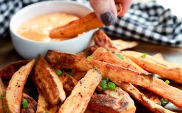 A stack of sweet potato fries on a plate with a woman's hand dipping a fry into a small white bowl of aioli.