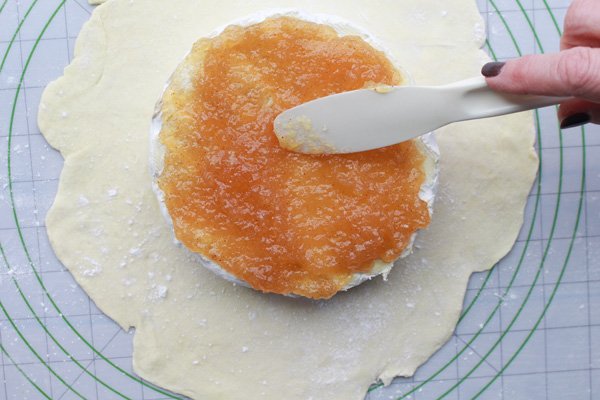 A person spreading ginger jam on top of a round brie placed on top of uncooked puff pastry.