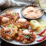 Golden-brown and crispy coconut shrimp cakes on a round white serving plate with a spicy aioli sauce, lemon wedges, and hot red peppers on this side and a glass of white wine in the background.